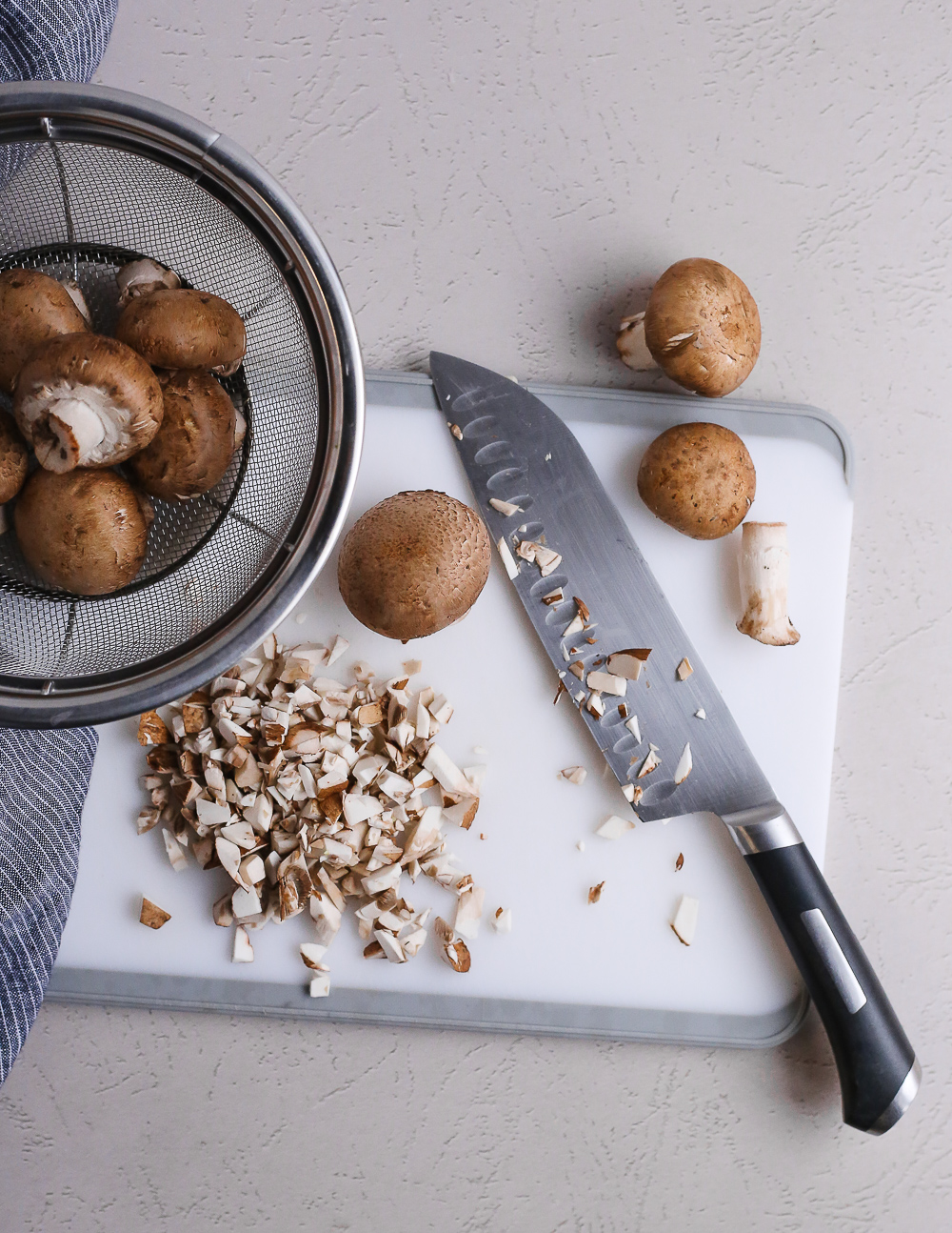 Overhead view of a cutting board and knife with whole and partially chopped mushrooms, indicating both the caps and stems of the mushrooms are included in the recipe