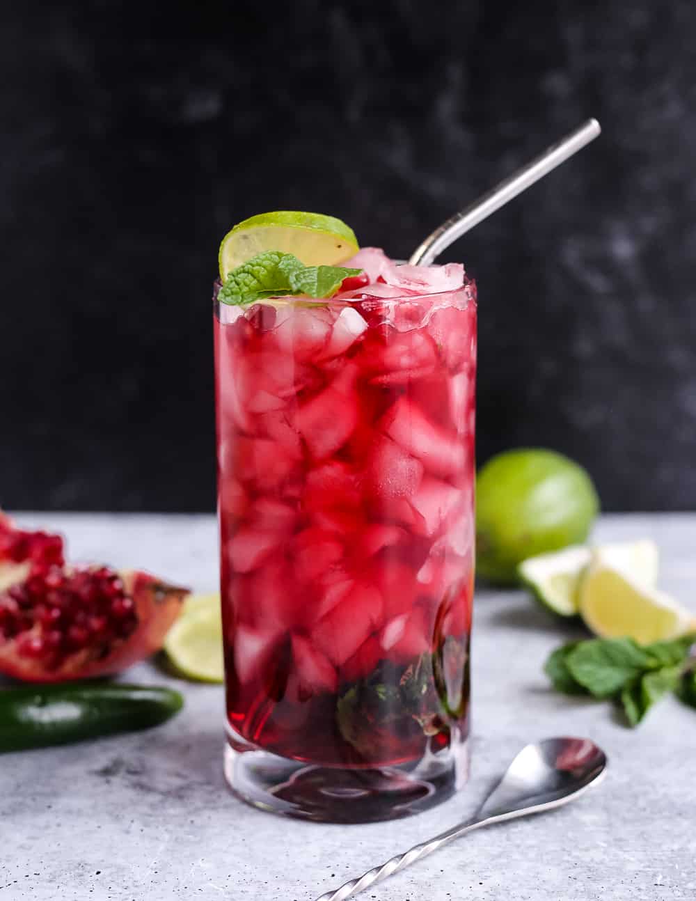 A bright red pomegranate mocktail on a countertop with lime wedge and mint garnish, served in a tall glass with a silver straw