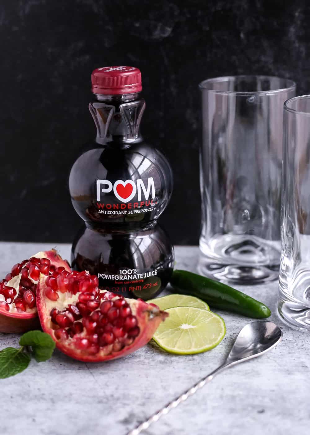 A bottle of pomegranate juice and part of a pomegranate on a kitchen countertop along with the other ingredients and a glass for a Spicy Pomegranate Mojito Mocktail