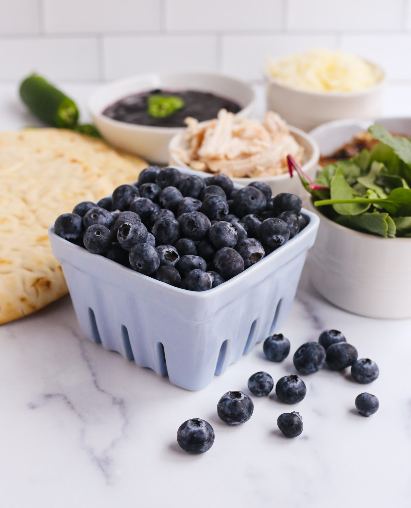 A light blue ceramic bowl holding fresh blueberries, along with other ingredients on a kitchen counter for chicken flatbread ideas for summer