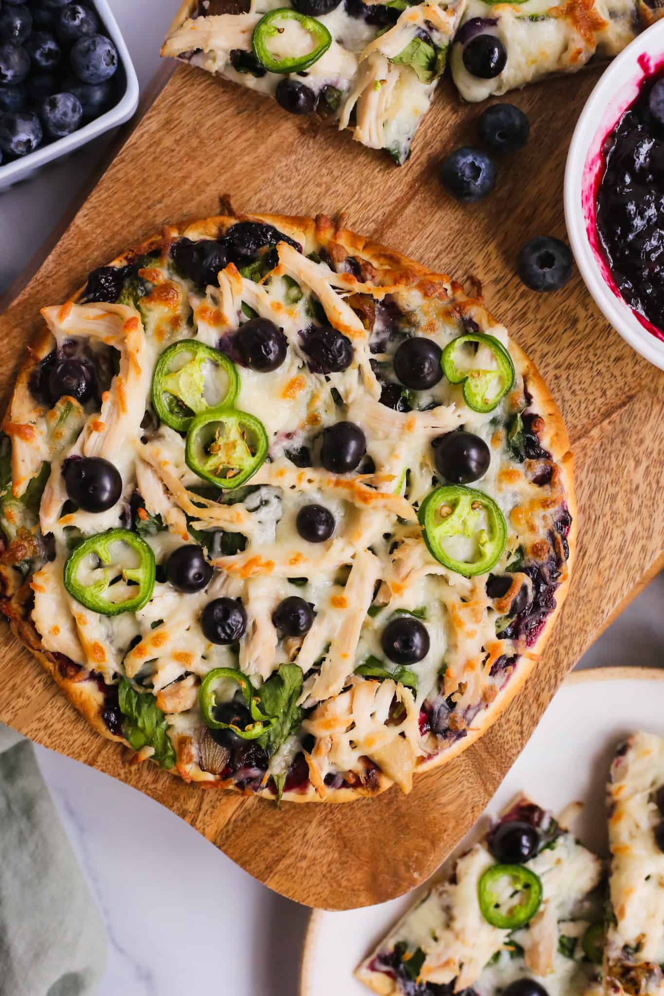 Overhead view of a chicken flatbread with blueberry balsamic sauce on a serving board, garnished with blueberries, sliced jalapeños, and melted cheese