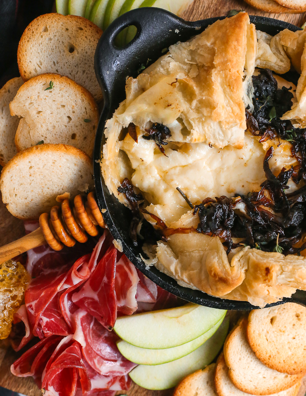 A close view into a baked brie appetizer wrapped in puff pastry and served on a charcuterie board. It's topped with dark caramelized onions and the cheese is gooey and melty