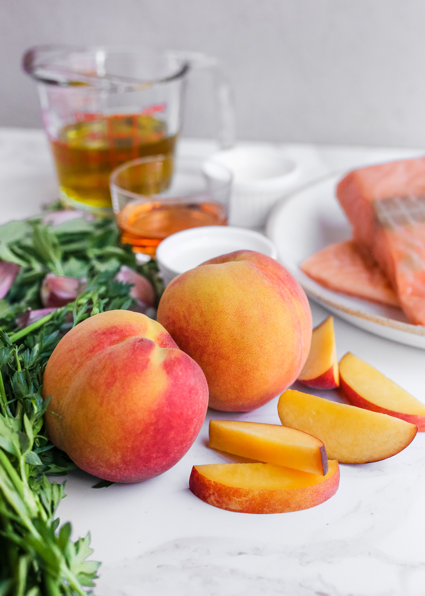 Two fresh peaches rest among other ingredients for a summer salmon salad, including fresh oregano, garlic cloves, red wine vinegar and olive oil, and fresh salmon fillets. A few slices of peaches are also including, showing the vibrant orange interior