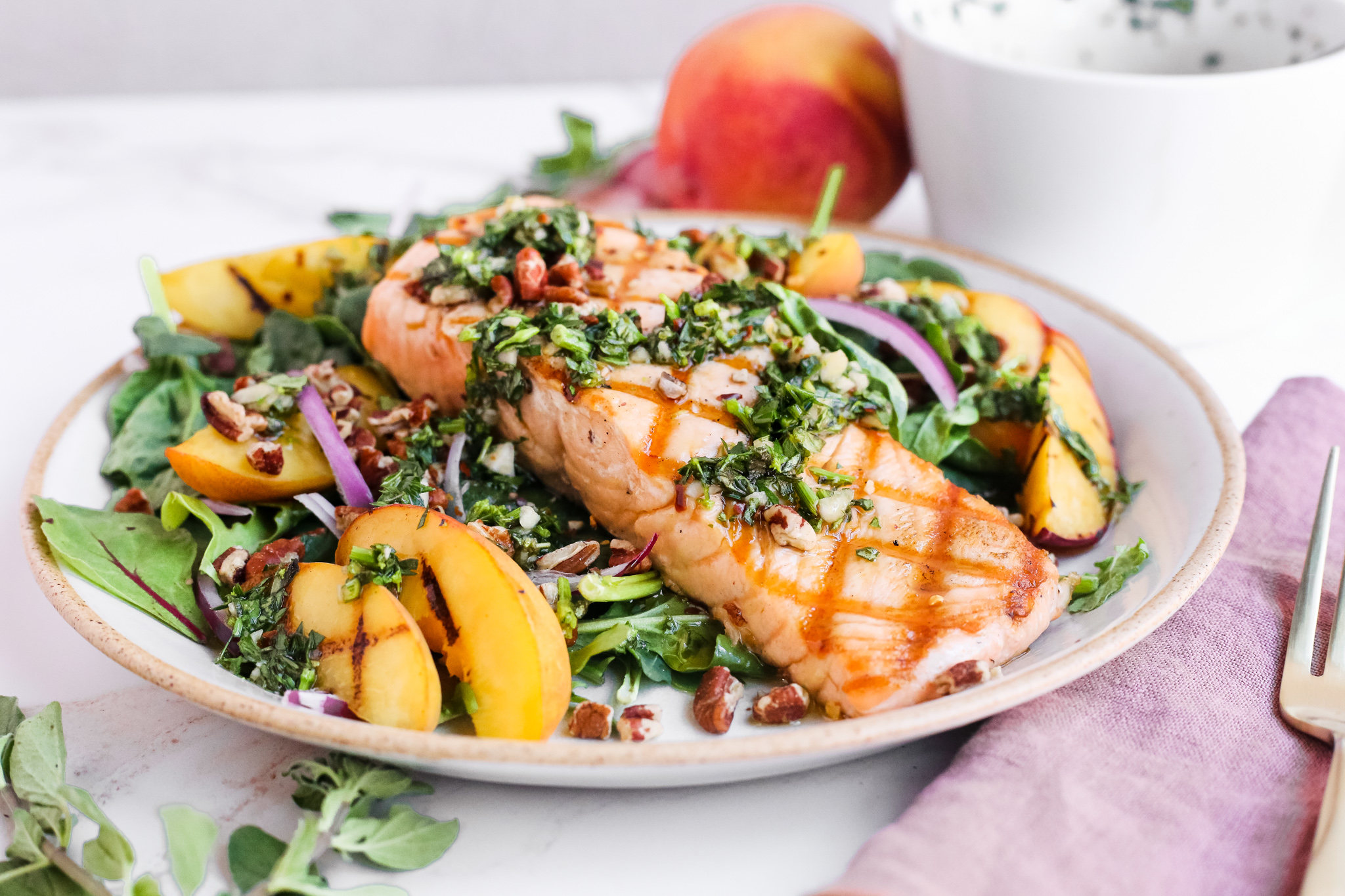 Landscape image of a seared piece of salmon with prominent grill marks, served on a bed of leafy salad greens with grilled peach slices, red onions, pecans, and a chunky chimichurri 
