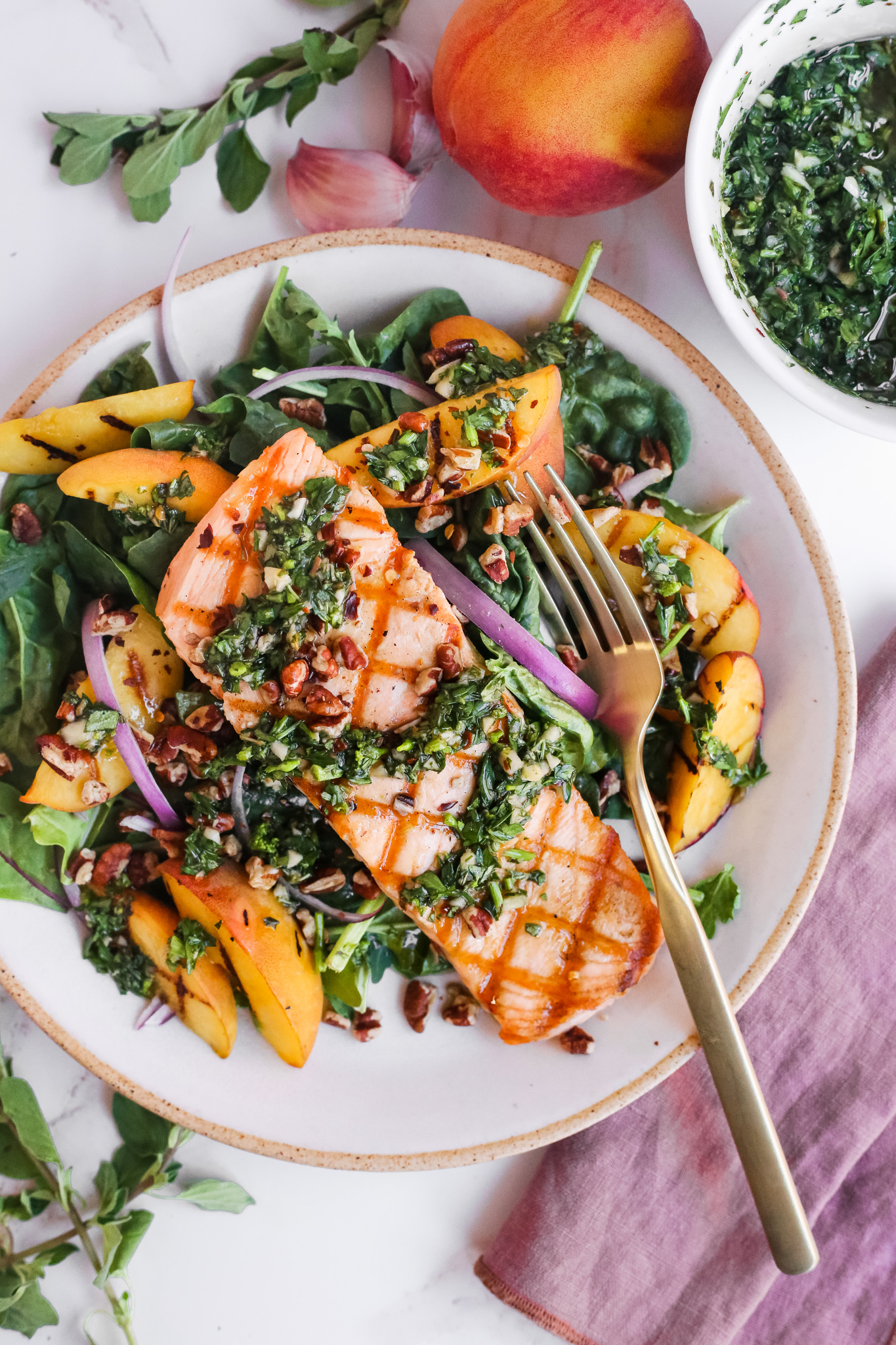 A colorful salad with grilled salmon, peach slices, red onions, walnuts, and a chunky Chimichurri dressing, on a beige ceramic plate with a brass colored fork placed on the side