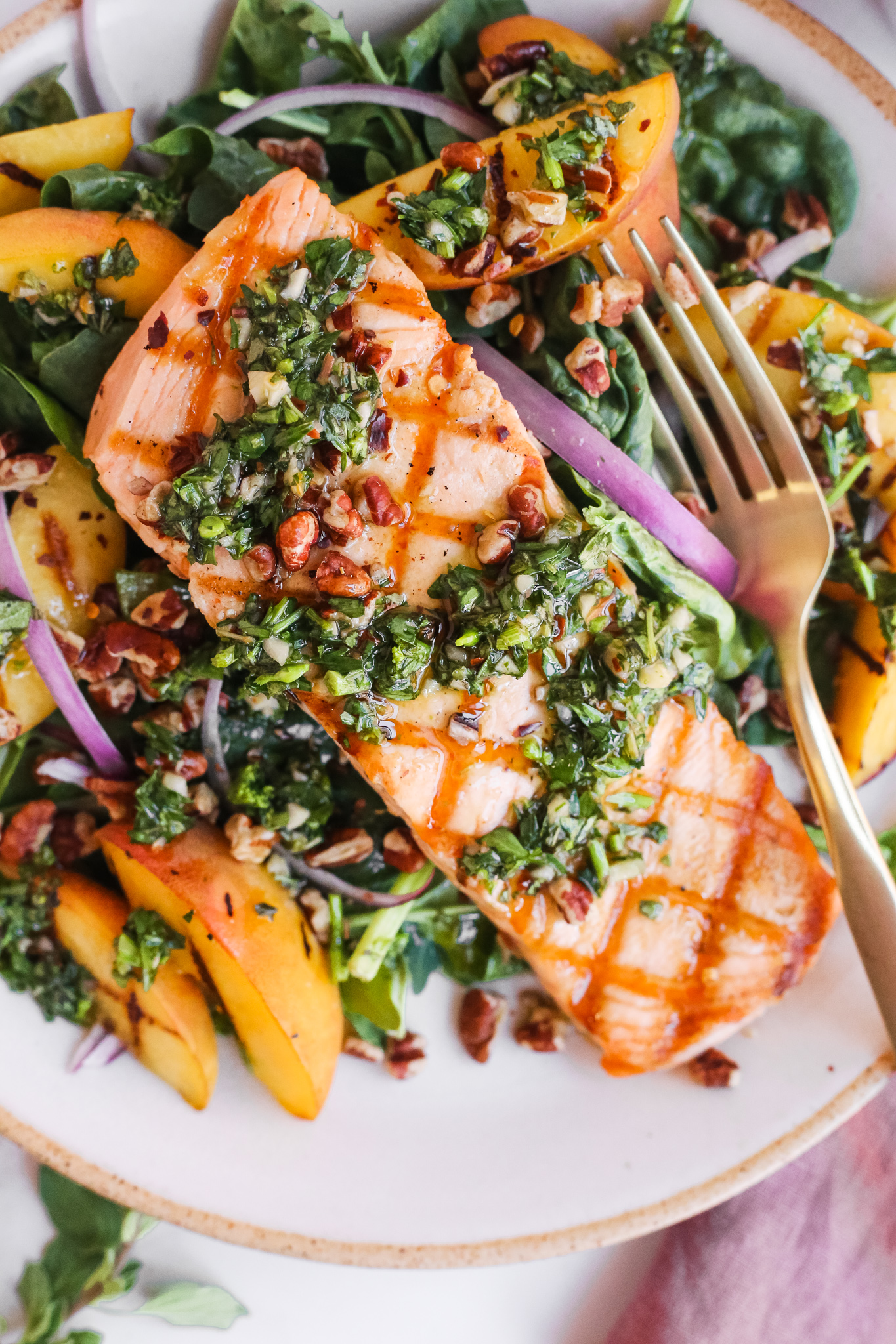 Closeup view of a summer salmon salad recipe with grilled peaches and chimichurri, served on a glazed ceramic plate with a gold fork
