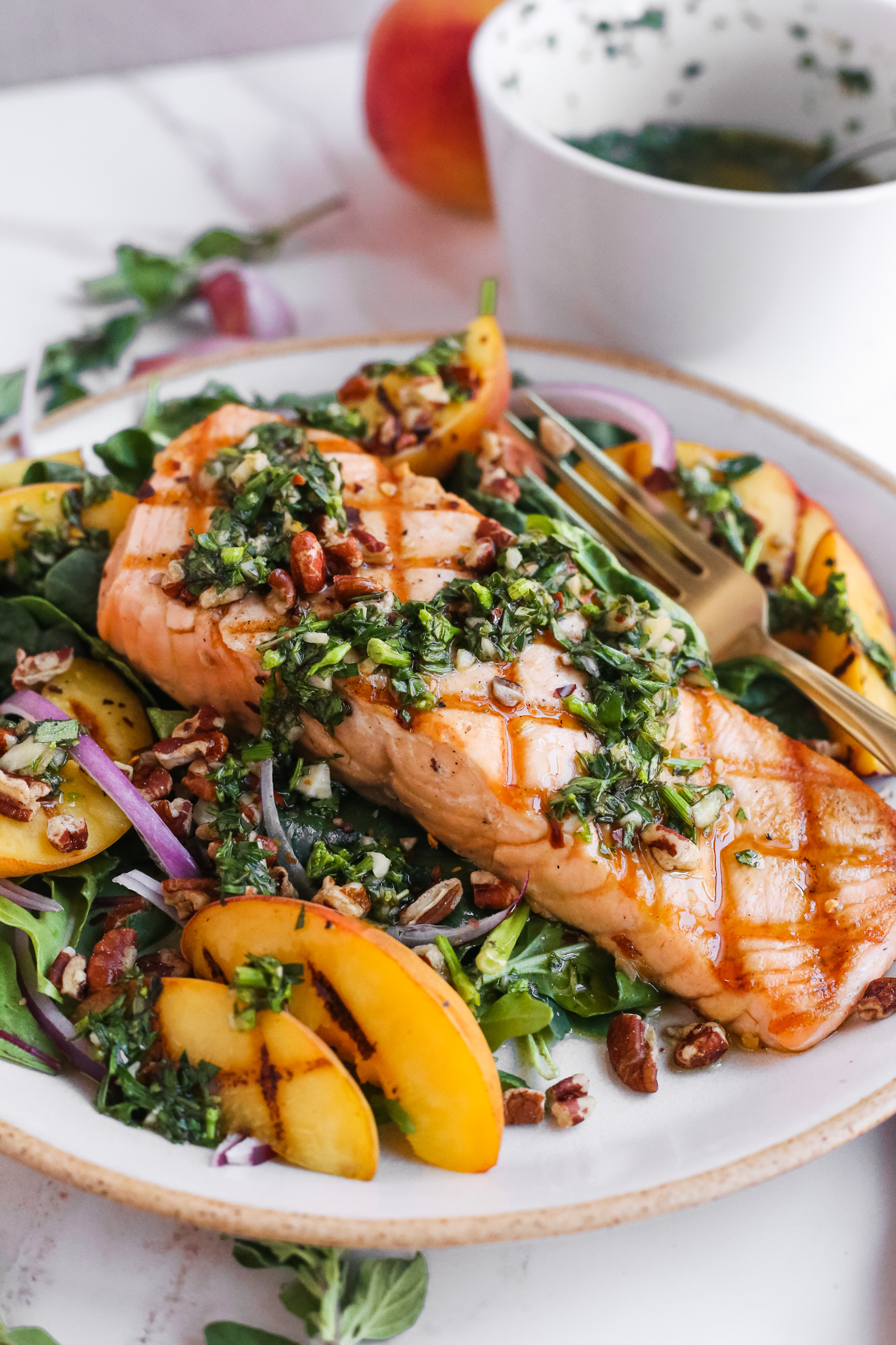 A glazed ceramic plate with a summer salmon salad with grilled peaches and chimichurri, served with extra chimichurri in the background