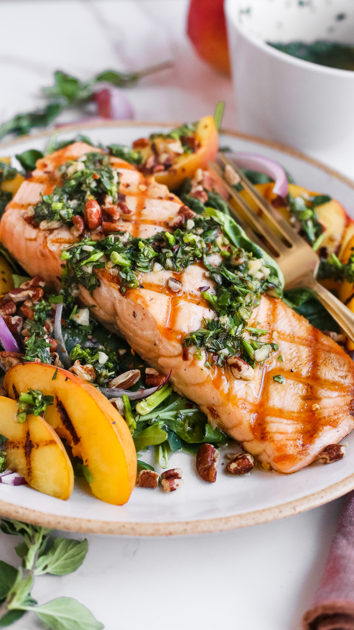 A close view of a grilled salmon fillet, served on a bed of salad greens and topped with grilled peaches, chopped pecans, sliced red onions, and a chimichurri sauce
