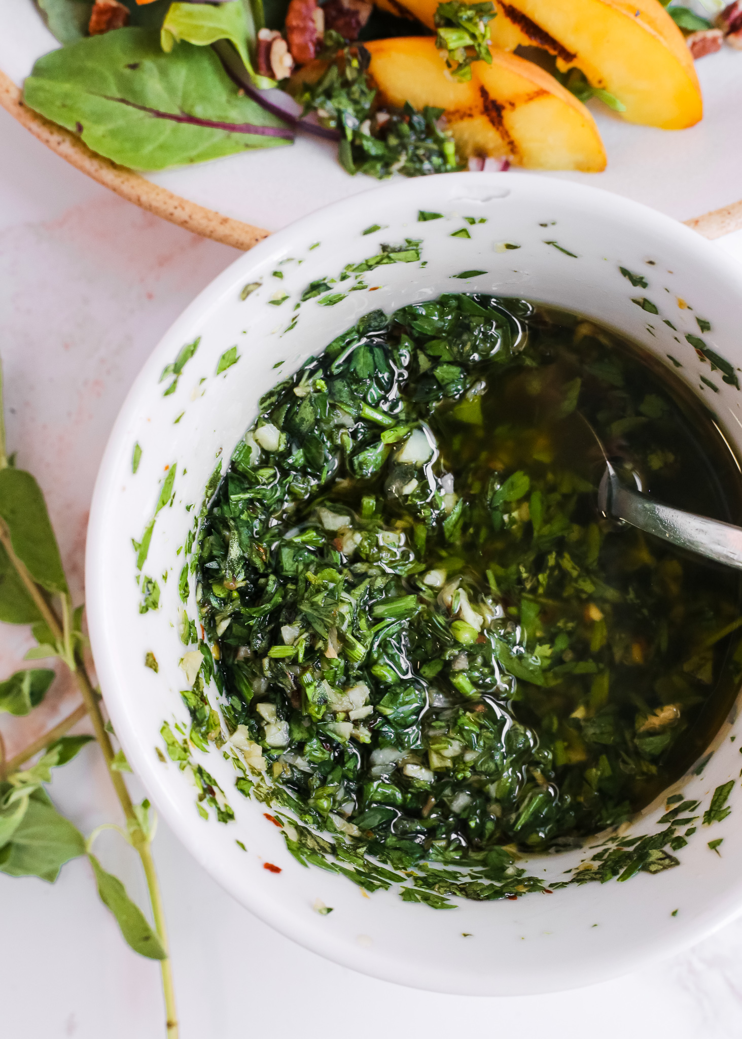 A white ceramic dish of chimichurri with parsley, garlic, oregano, red pepper flakes, olive oil, and red wine vinegar, pictured with a summer salmon salad in the background