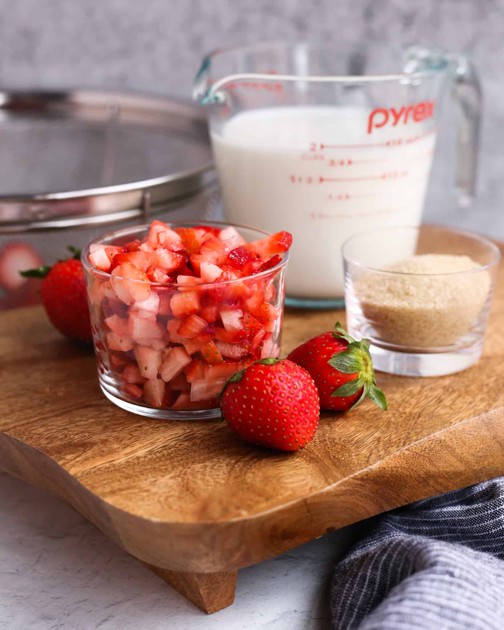 Roughly chopped fresh strawberries in a glass ramekin, shown on a wooden tray with fresh strawberries, turbinado sugar, and whole milk in a clear glass measuring cup