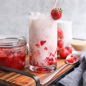A glass of Korean strawberry milk served on a wooden tray with strawberry puree, fresh strawberries, and whole milk in the background