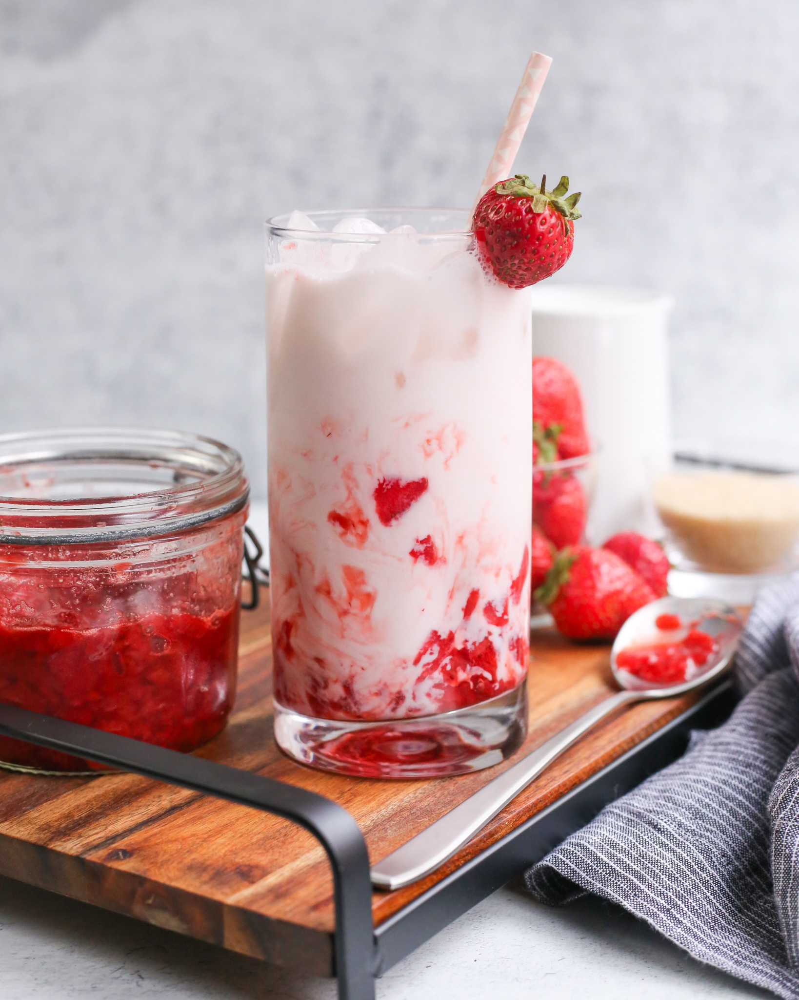 A glass of Korean Strawberry Milk is served on a wooden tray along with a strawberry puree and fresh strawberries. A container of whole milk is seen in the background