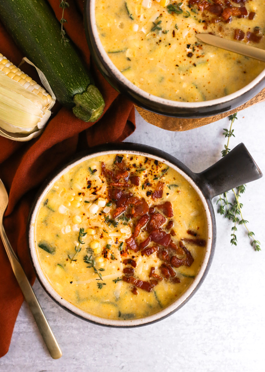 Overhead view of a creamy corn chowder recipe in black and beige ceramic bowls with a handle, garnished with bacon, cheese, and fresh thyme