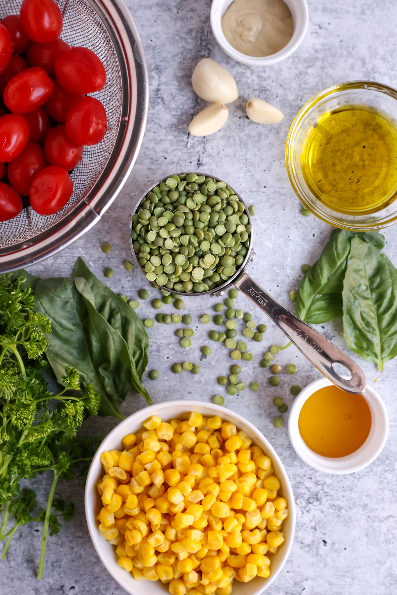 Arrangements on ingredients on a kitchen countertop, including uncooked green split peas, cheery tomatoes, garlic cloves, olive oil, fresh basil and parsley, and sweet corn
