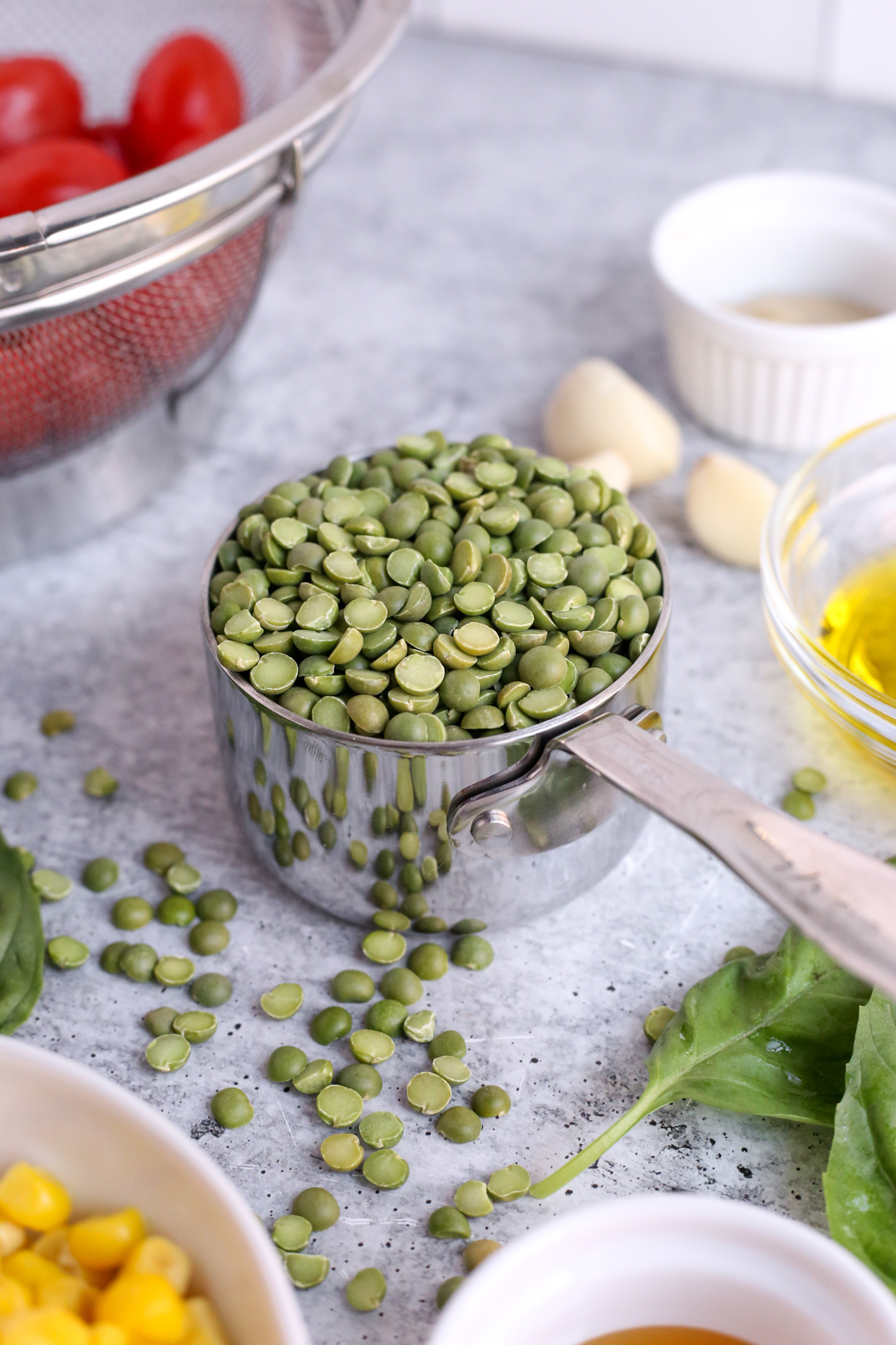 A close up shot of a metal measuring cup with uncooked green split peas, surrounded by other ingredients for summer split pea salad recipe