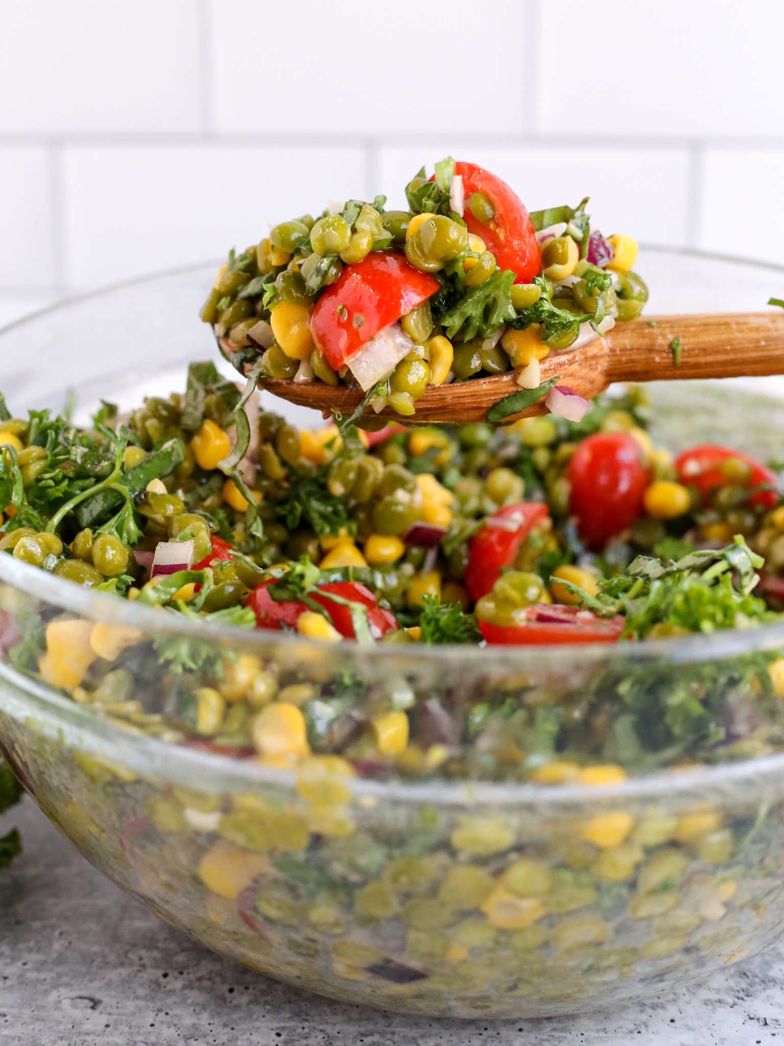 A wooden serving spoon holds up and displays a scoop of a summer split pea salad, served out of a clear glass mixing bowl against a white kitchen background