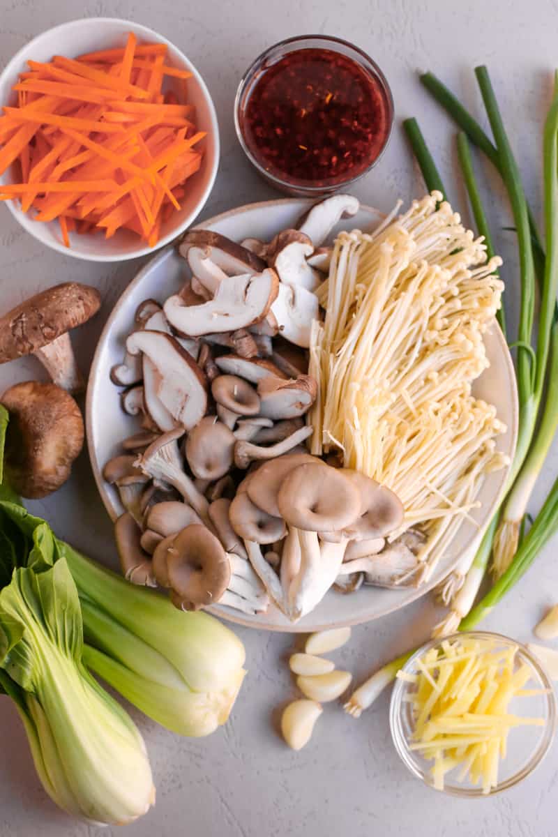 Ingredients for a simple mushroom stir fry recipe arranged on a kitchen counter, including a variety of mushroom in the center, thinly sliced carrots and ginger, baby wok chop, green onions, and a clear glass ramekin of chili oil