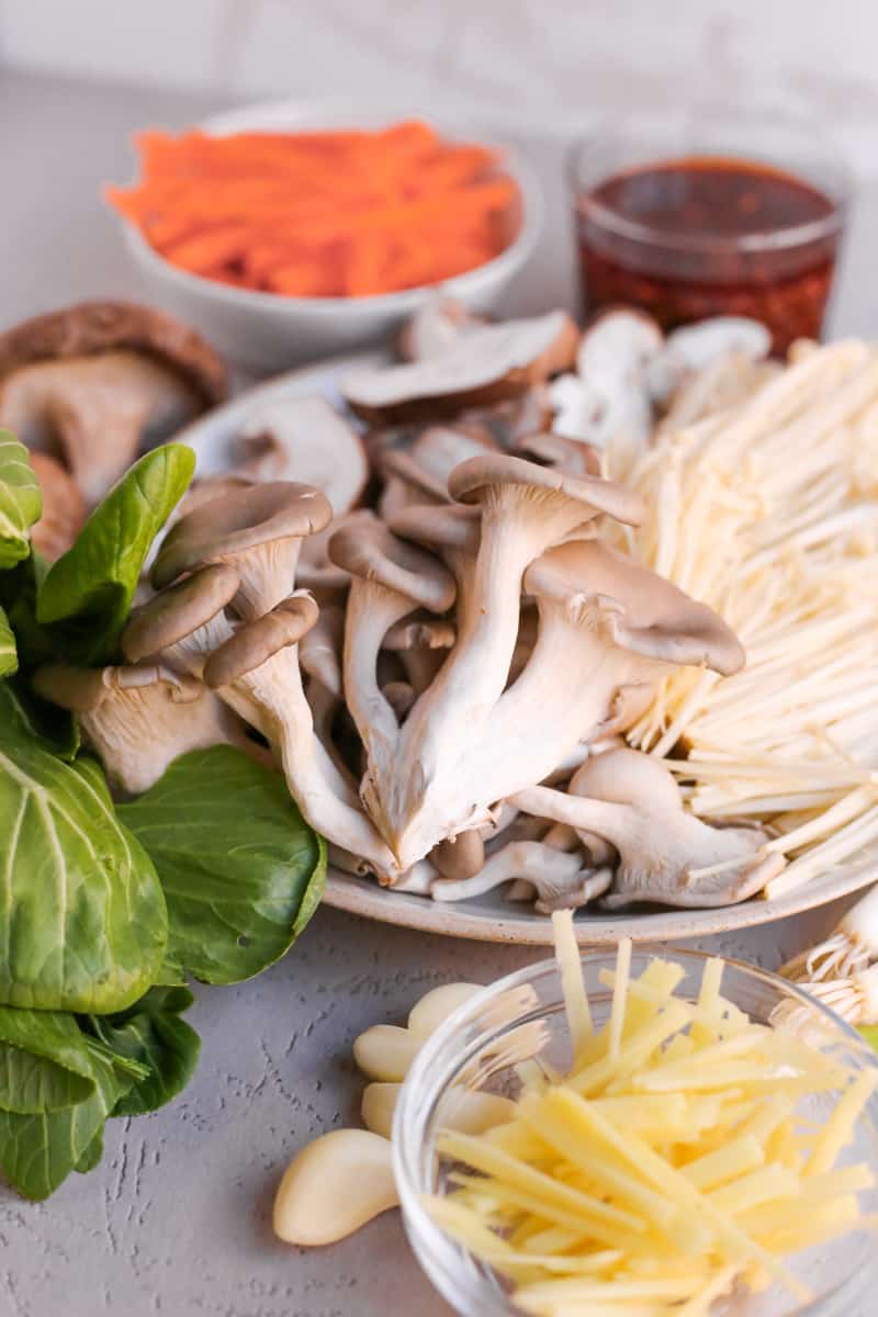 Ingredients for a simple mushroom stir fry recipe arranged on a kitchen counter, including a variety of mushroom in the center, thinly sliced carrots and ginger, baby wok chop, green onions, and a clear glass ramekin of chili oil