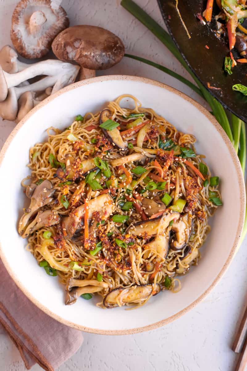 Overhead view of a bowl full of stir fried mushrooms and ginger garlic noodles, surrounded by a wok, extra mushrooms, green onions, and a pink linen