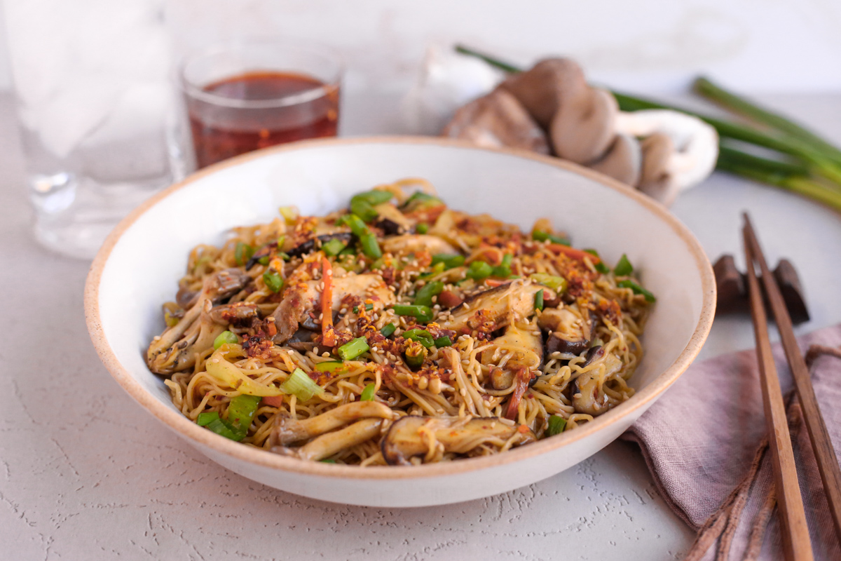 Ceramic bowl filled with mushroom stir ry noodles and garnished with green onions, chili oil, and sesame seeds, served with dark wooden chopsticks and a glass of ice water