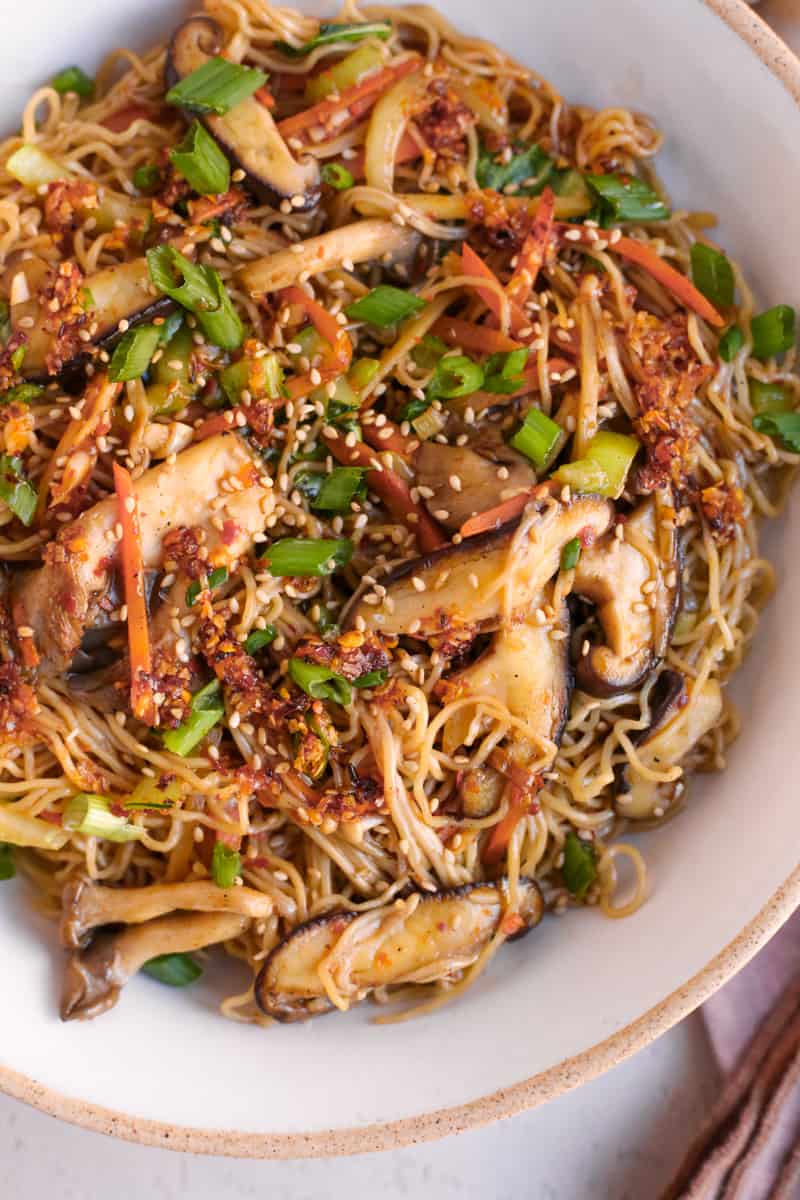 Closer view of mushroom stir fry noodles in a serving bowl, garnished with green onions, toasted sesame seeds, and chili oil