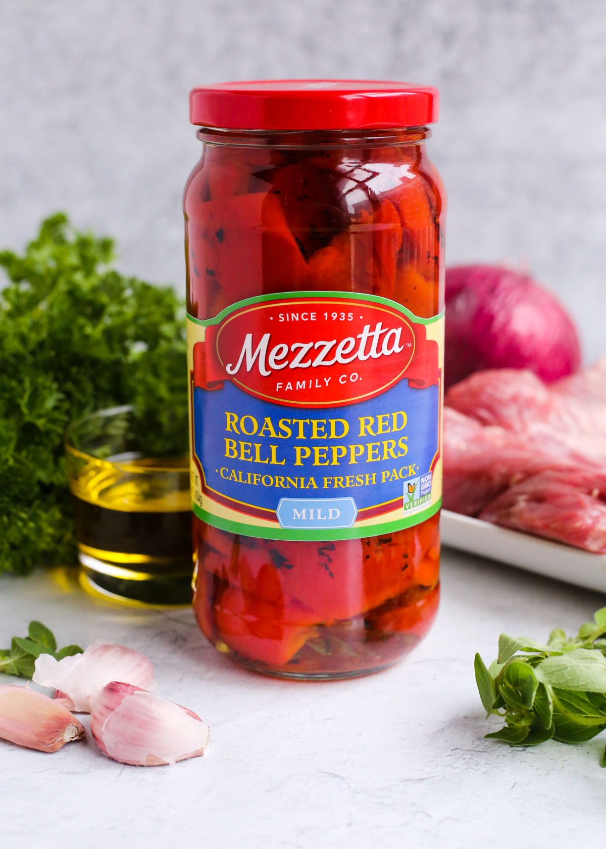 An unopened glass jar of Mezzetta roasted red peppers on a kitchen countertop, surrounded by a clear glass ramekin of olive oil, garlic cloves, green herbs, and a red onion