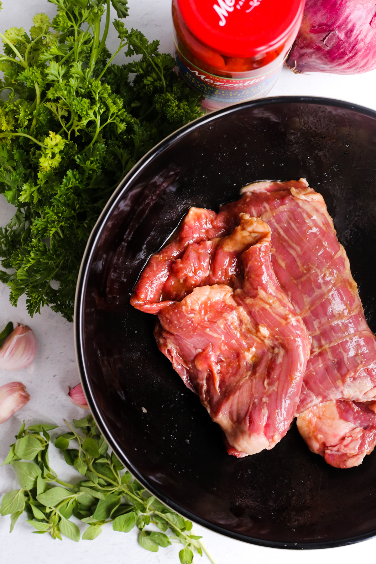 Overhead view of a marinated skirt steak in a black ceramic mixing bowl, surrounded by green herbs, garlic cloves, a red onion, and jar of roasted red peppers