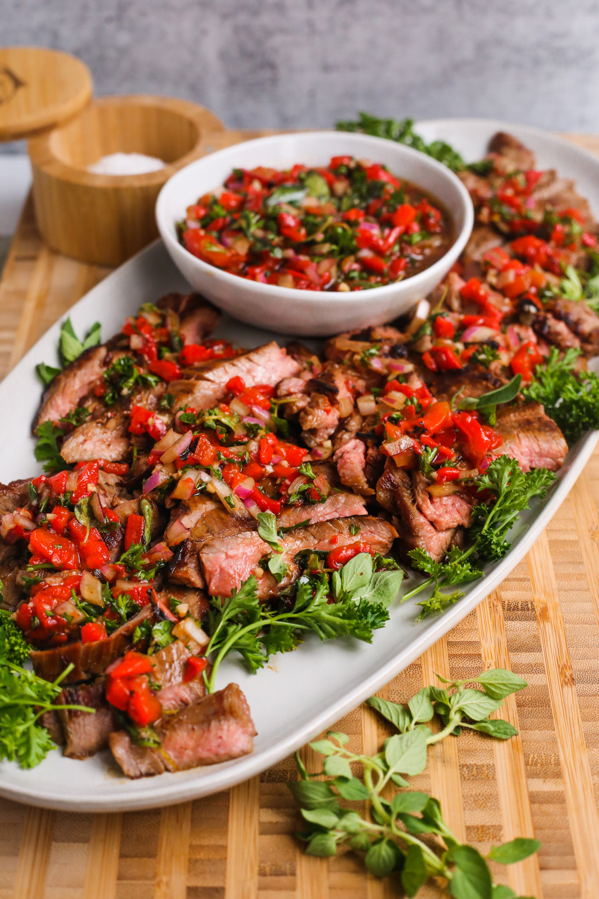 Side view of the skirt steak with roasted red pepper relish recipe, serving on a platter on top of a butcher block board with a salt cellar and fresh herbs in the background