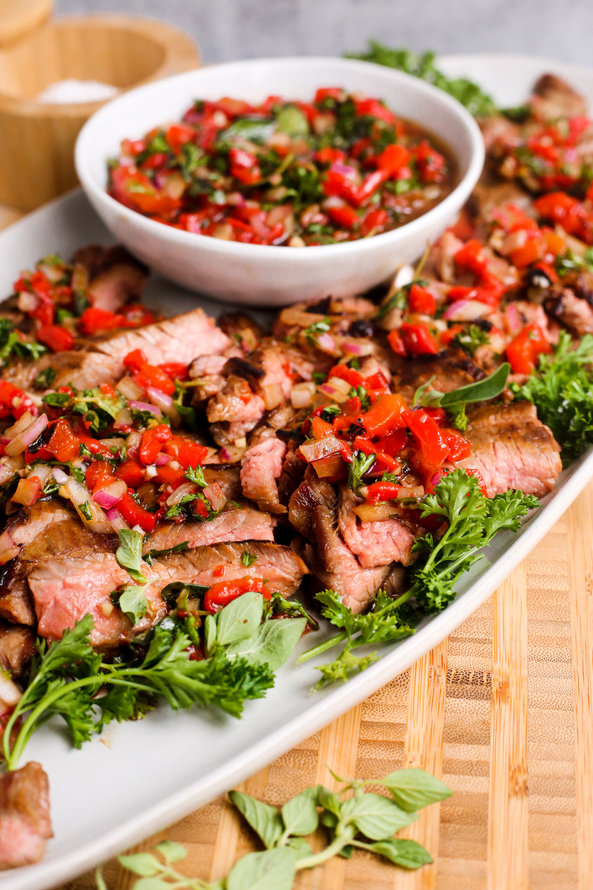 Side view of seared skirt steak, sliced against the grain, on a serving platter garnished with green herbs and a roasted red pepper relish