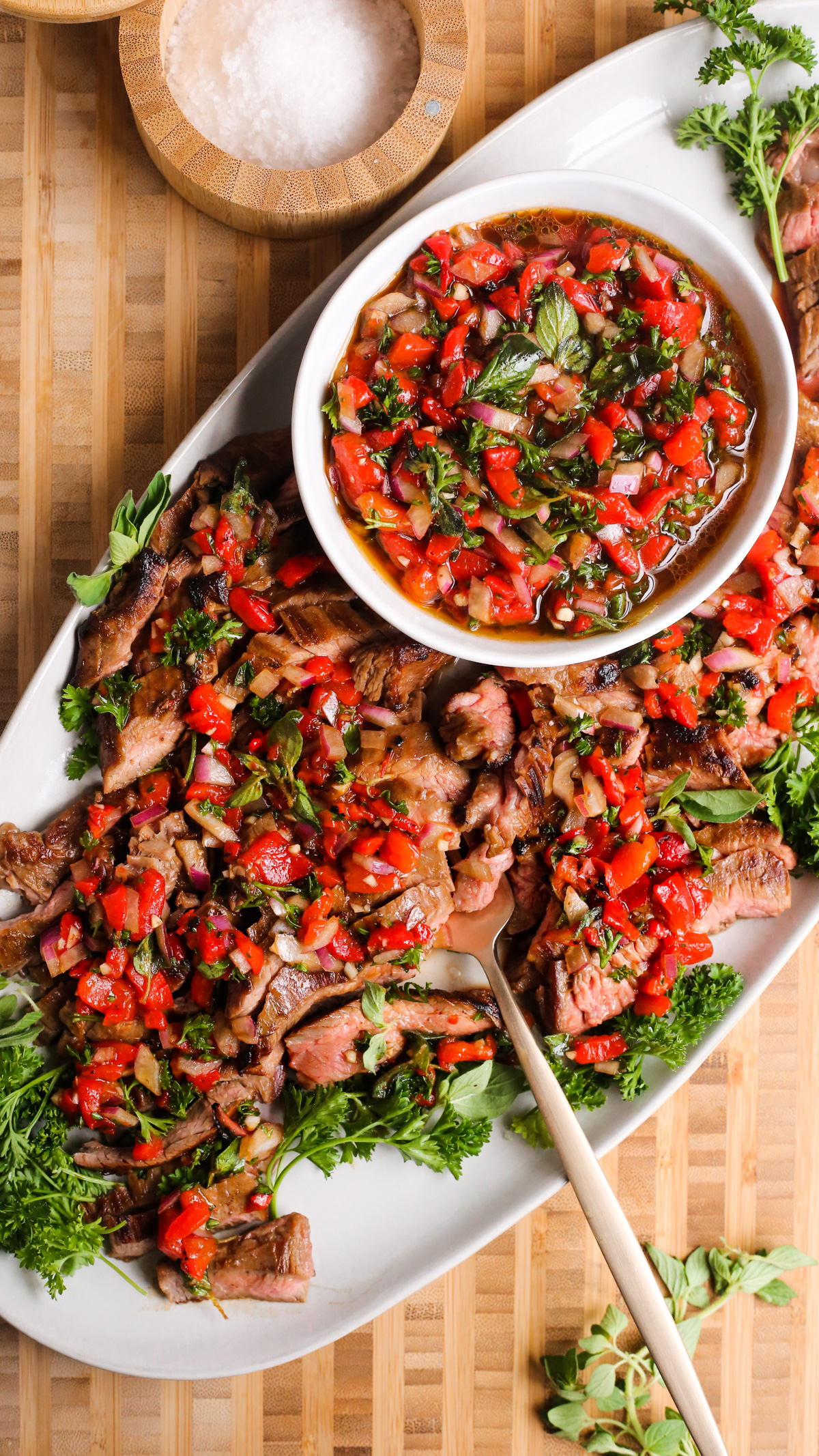 A serving platter of seared skirt steak with roasted red pepper relished, served on a butcher block board with a salt cellar and garnished with fresh herbs