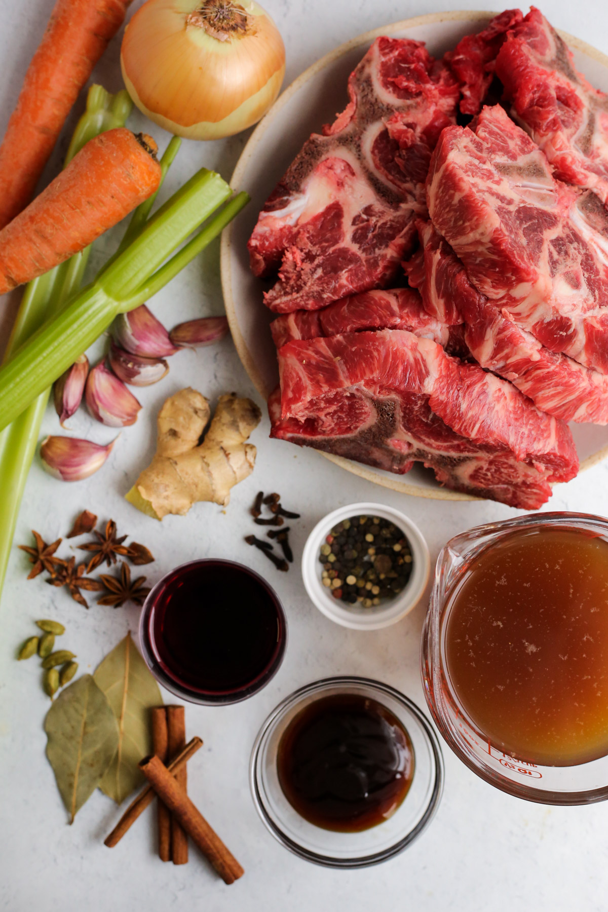 Overhead view of ingredients needed for a braised beef recipe, include beef neck bones, onion, carrots, celery, ginger, garlic, beef broth, and an assortment of dried spices and seasonings