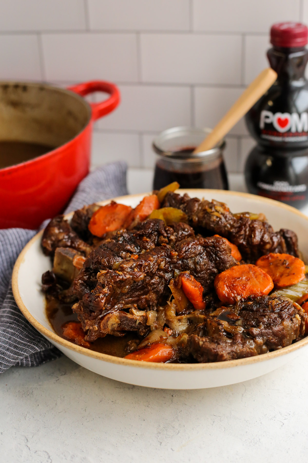 Plated bowl of braised beef neck bones in a seasoning braising liquid, served in front of a red dutch oven, bottle of pomegranate juice, and small glass jar of pomegranate glaze on a kitchen countertop