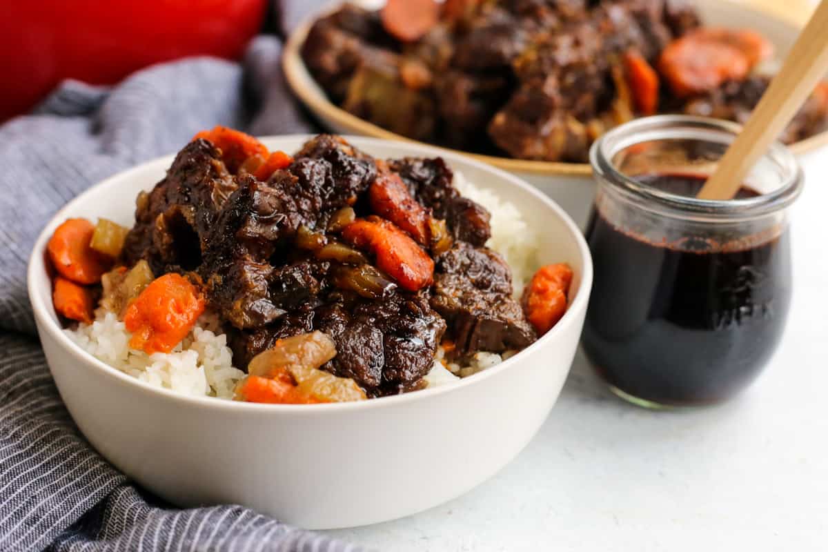 A white ceramic bowl full of braised beef, carrots, onions, and white rice next to a small glass jar of pomegranate glaze