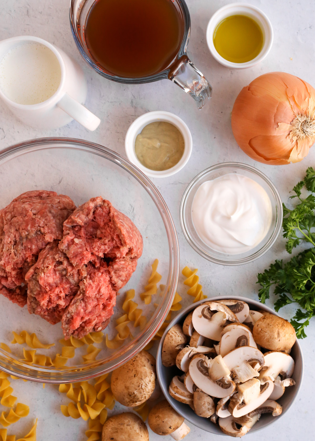 Display of ingredients to make a ground beef stroganoff recipe, include an onion, mushrooms, egg noodles, fresh parsley, various ramekins with sour cream, mustard, and beef broth, plus a prep bowl with uncooked ground beef