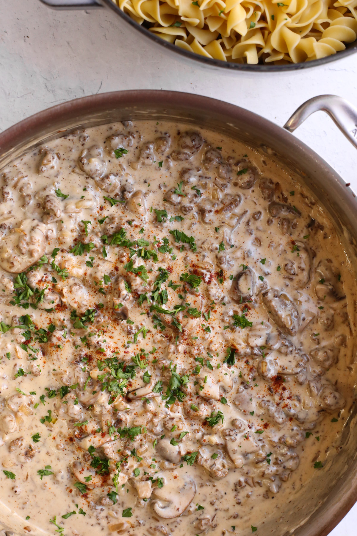 Overhead image of the large skillet filled with the ground beef stroganoff recipe, with a serving pot of egg noodles pictured above before serving