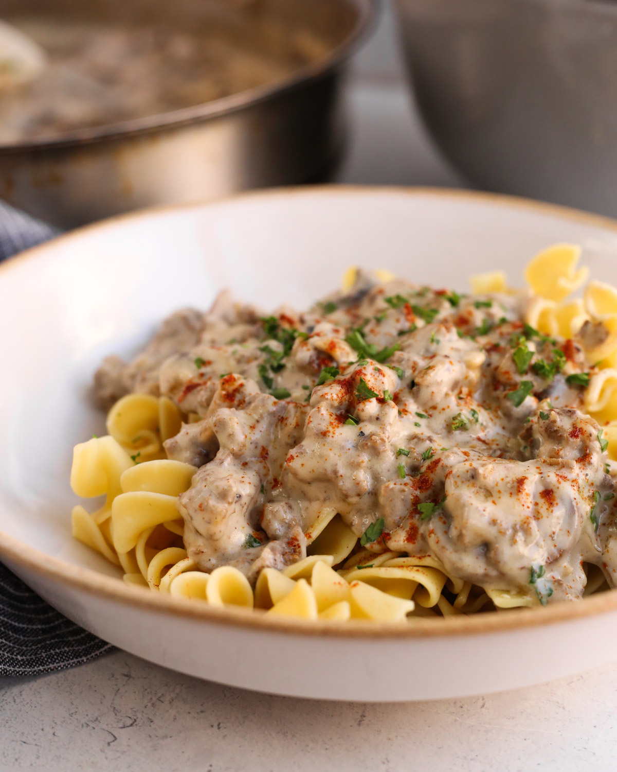 View into a large ceramic serving bowl filled with cooked egg noodles topped with a creamy mushroom gravy with bits of ground beef visible, garnished with parsley and paprika