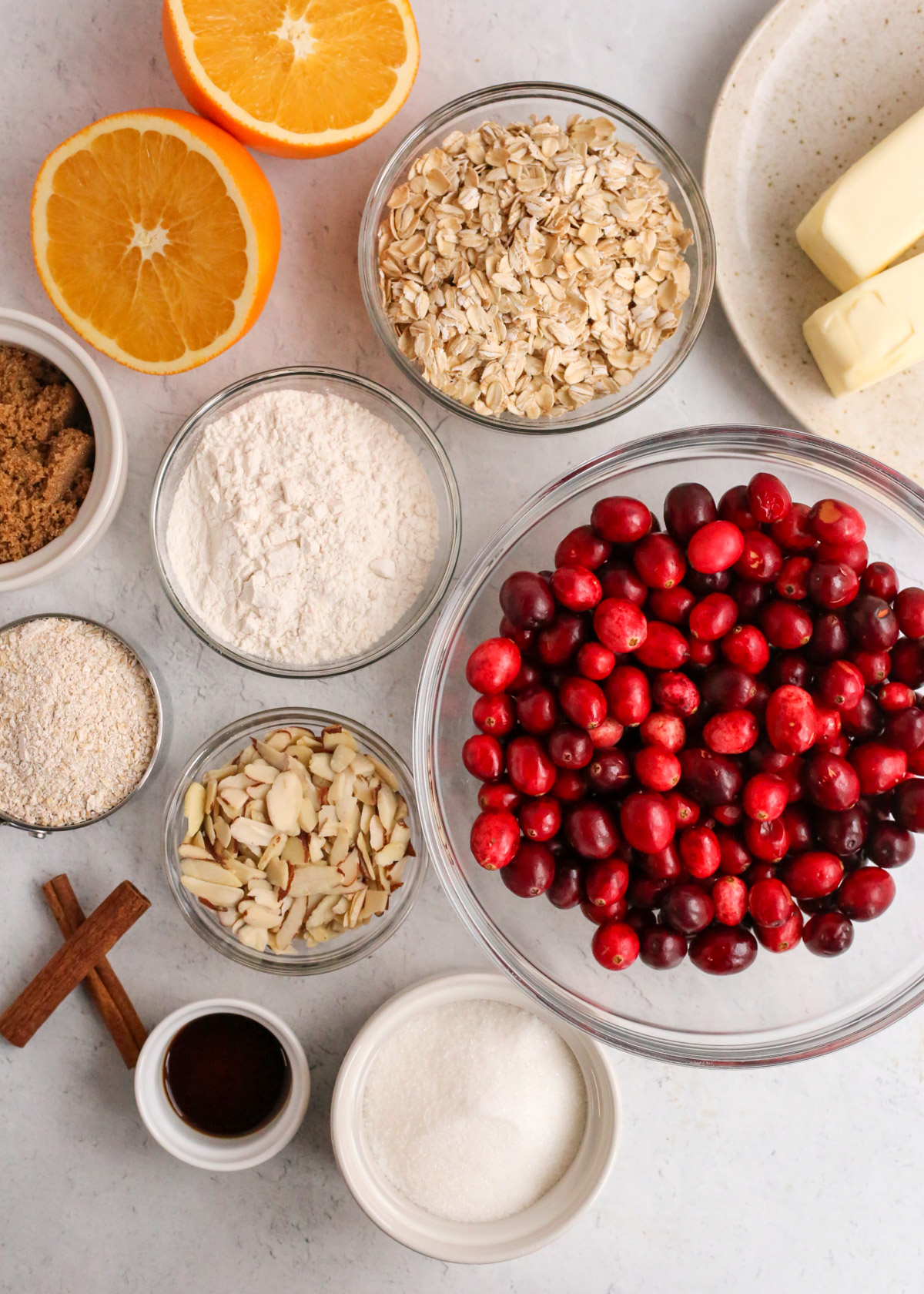 a collection of ingredients of an orange cranberry oat bar recipe, including fresh cranberries, sticks of butter, oats, flour, almonds, vanilla, brown and white sugar, and cinnamon sticks
