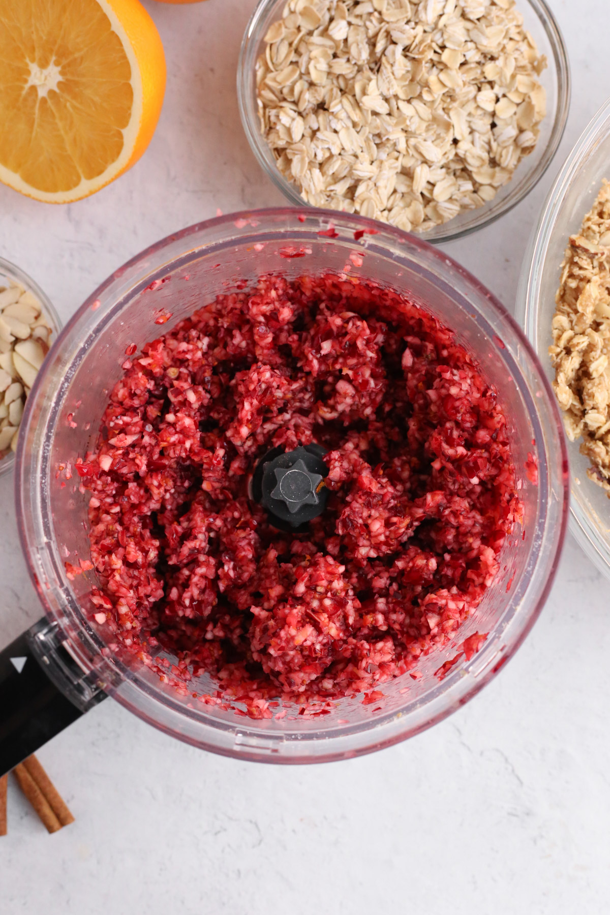 Overhead view into the bowl of a food processor that was used to chop fresh cranberries, with an orange, oats, and cinnamon sticks arranged around it