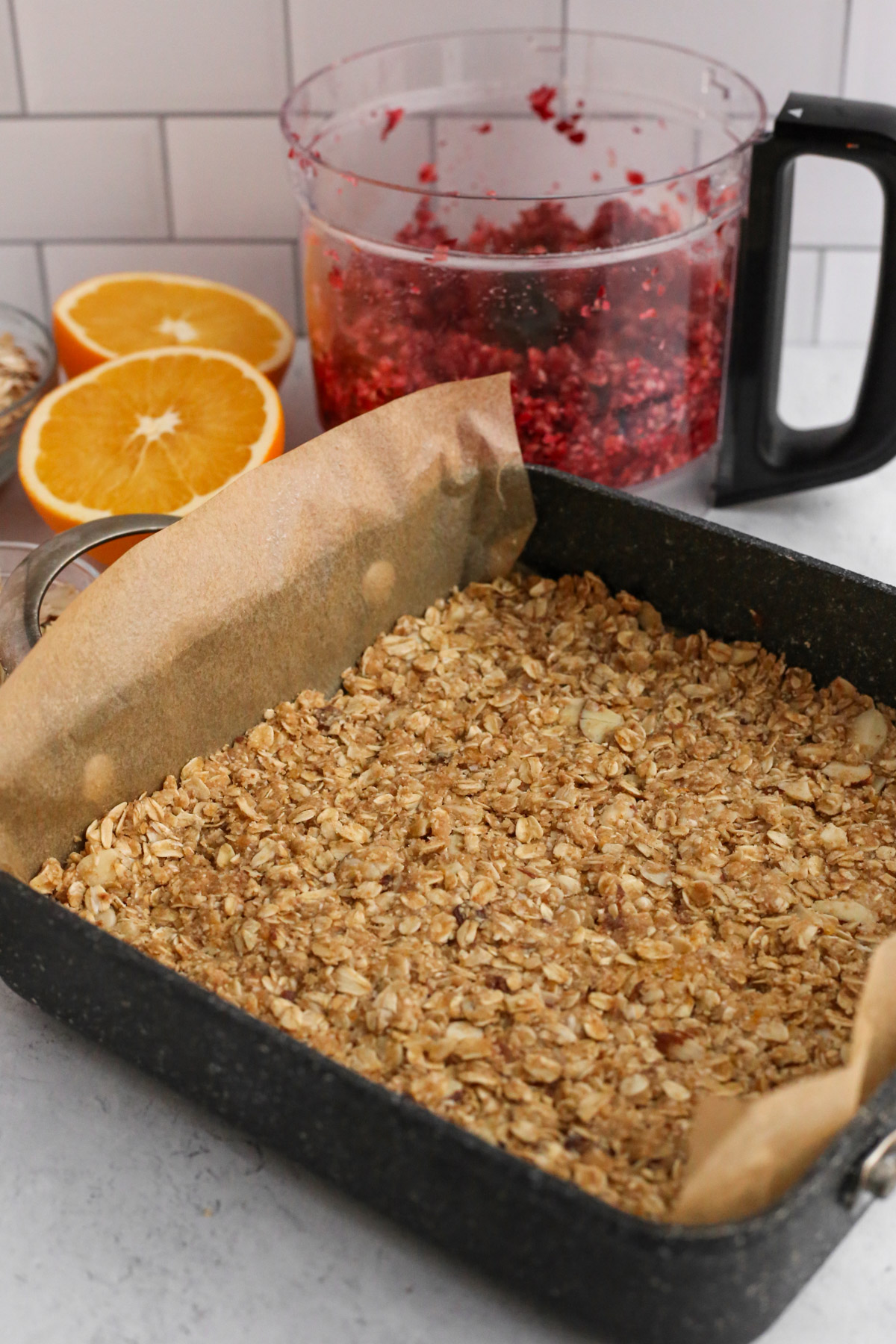 A parchment paper-lined square baking dish with a layer of crumbly oats pressed into a base layer.