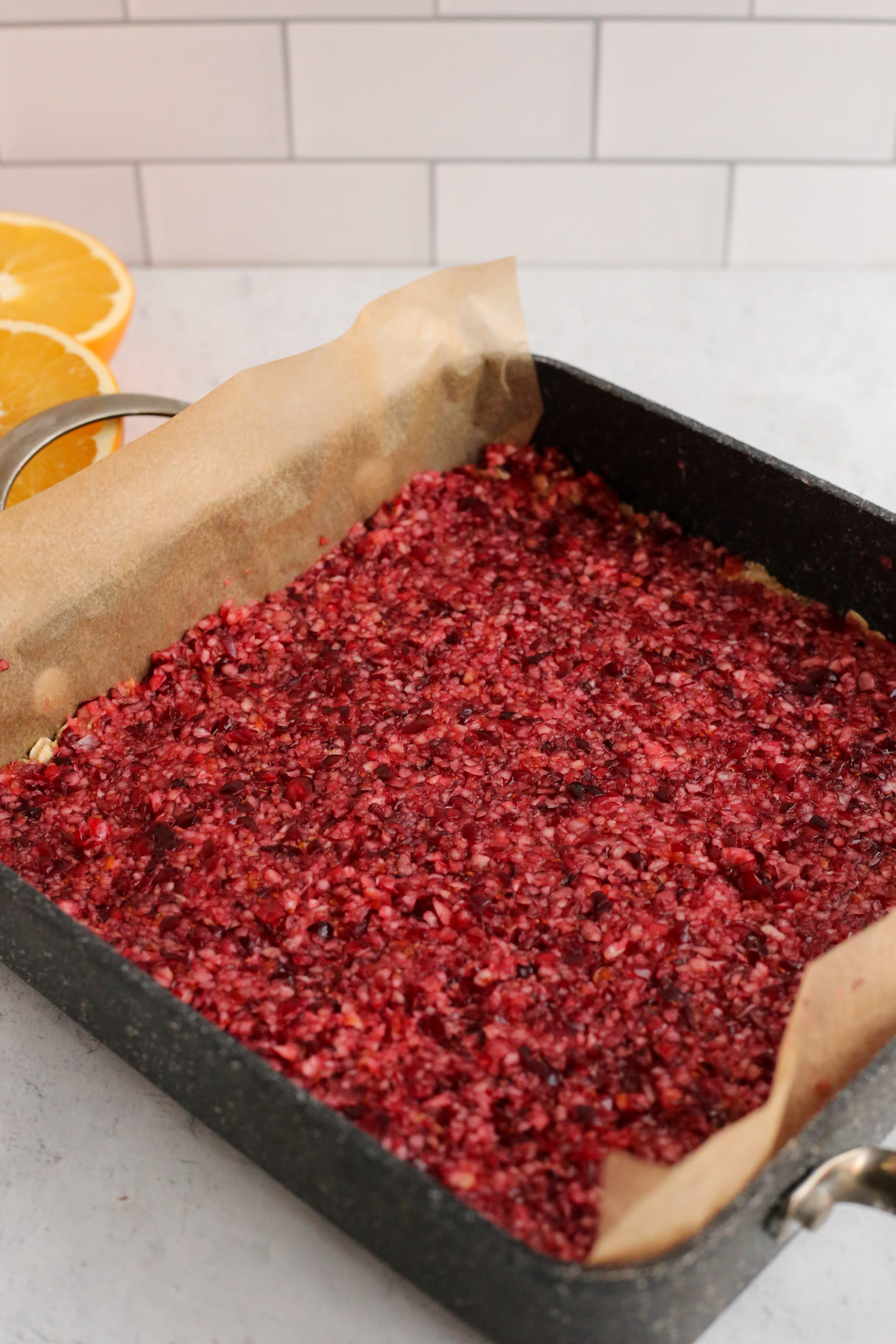 A square baking dish covered with a layer of chopped cranberries and orange zest, with sliced oranges in the background.