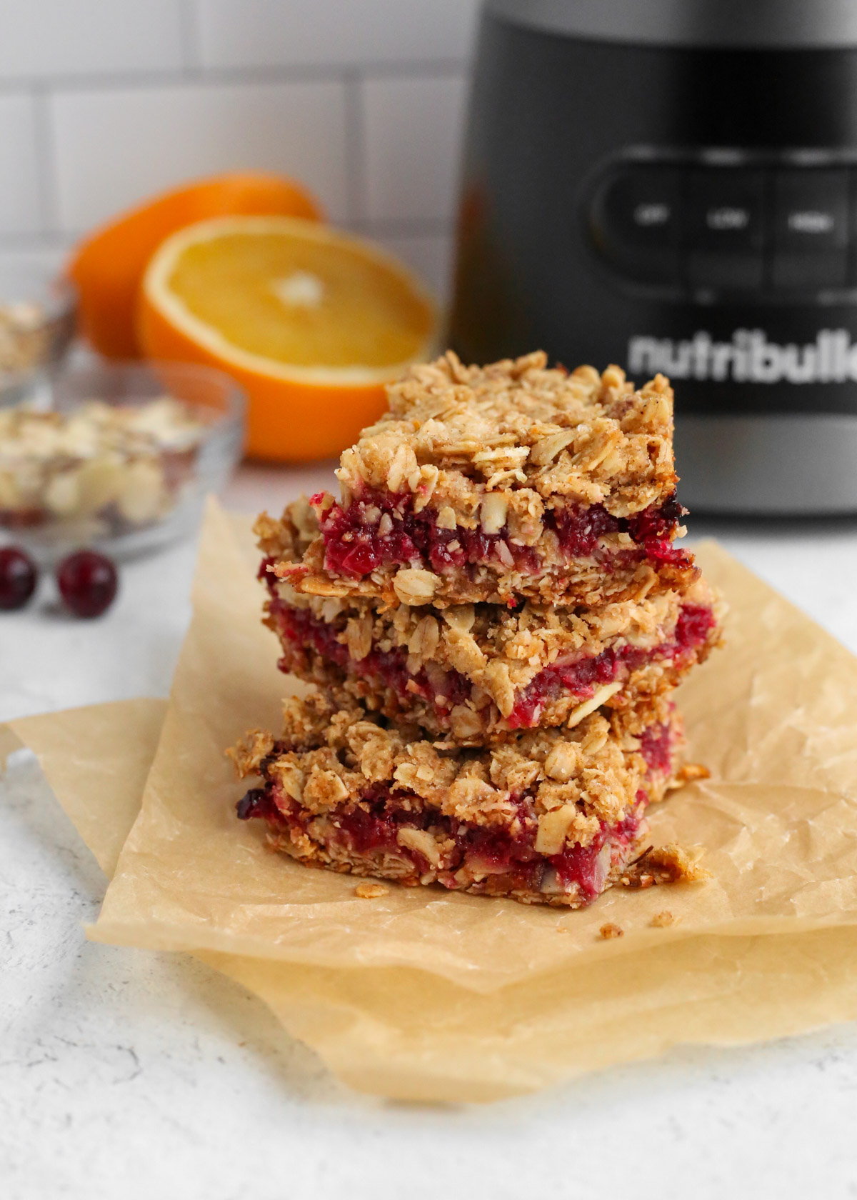 A stack of three orange cranberry oat bars on parchment paper, arranged in front of a food processor, sliced oranges, and loose cranberries