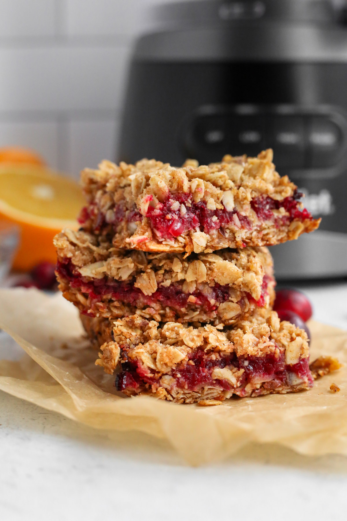 A close side shot of three orange cranberry oat bars, stacked on top of parchment paper in front of a food processor on a kitchen countertop