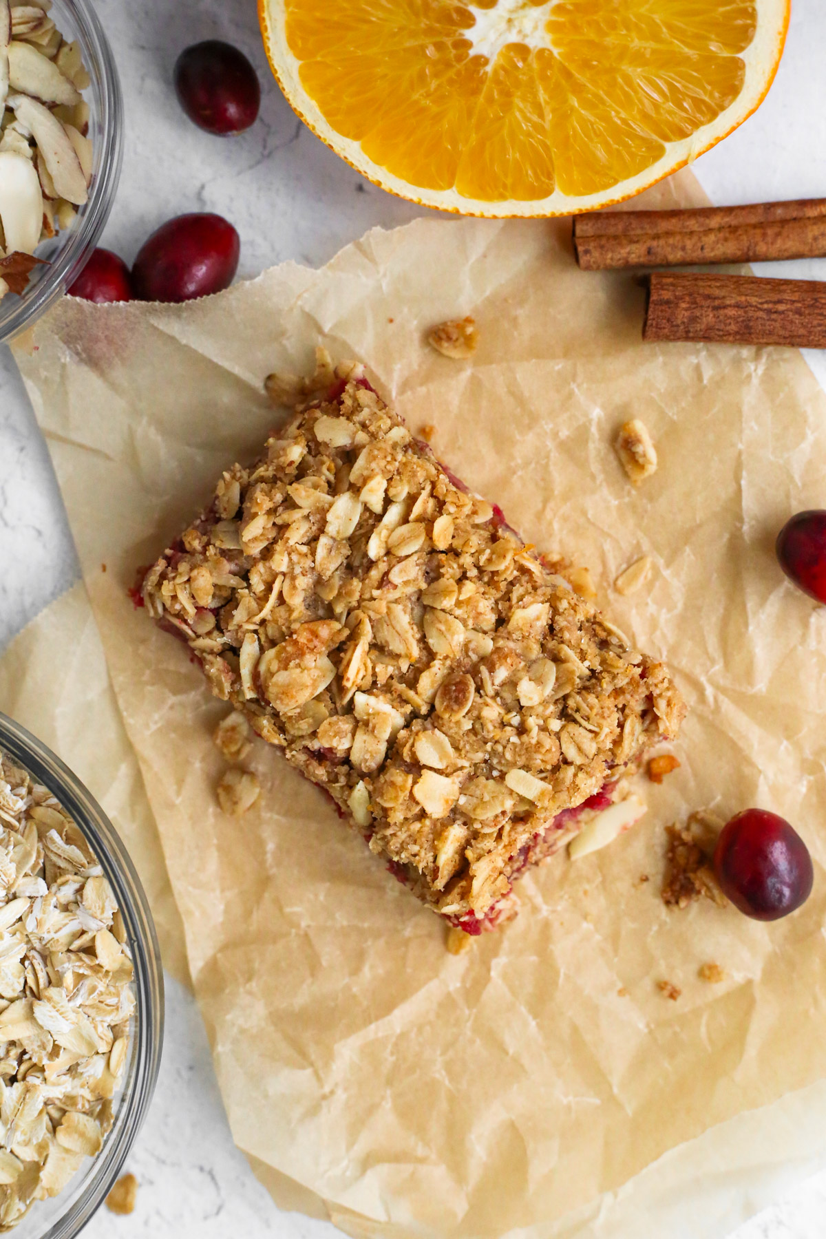 A single orange cranberry oat bar on parchment paper, surrounded by some of the other ingredients for the recipe
