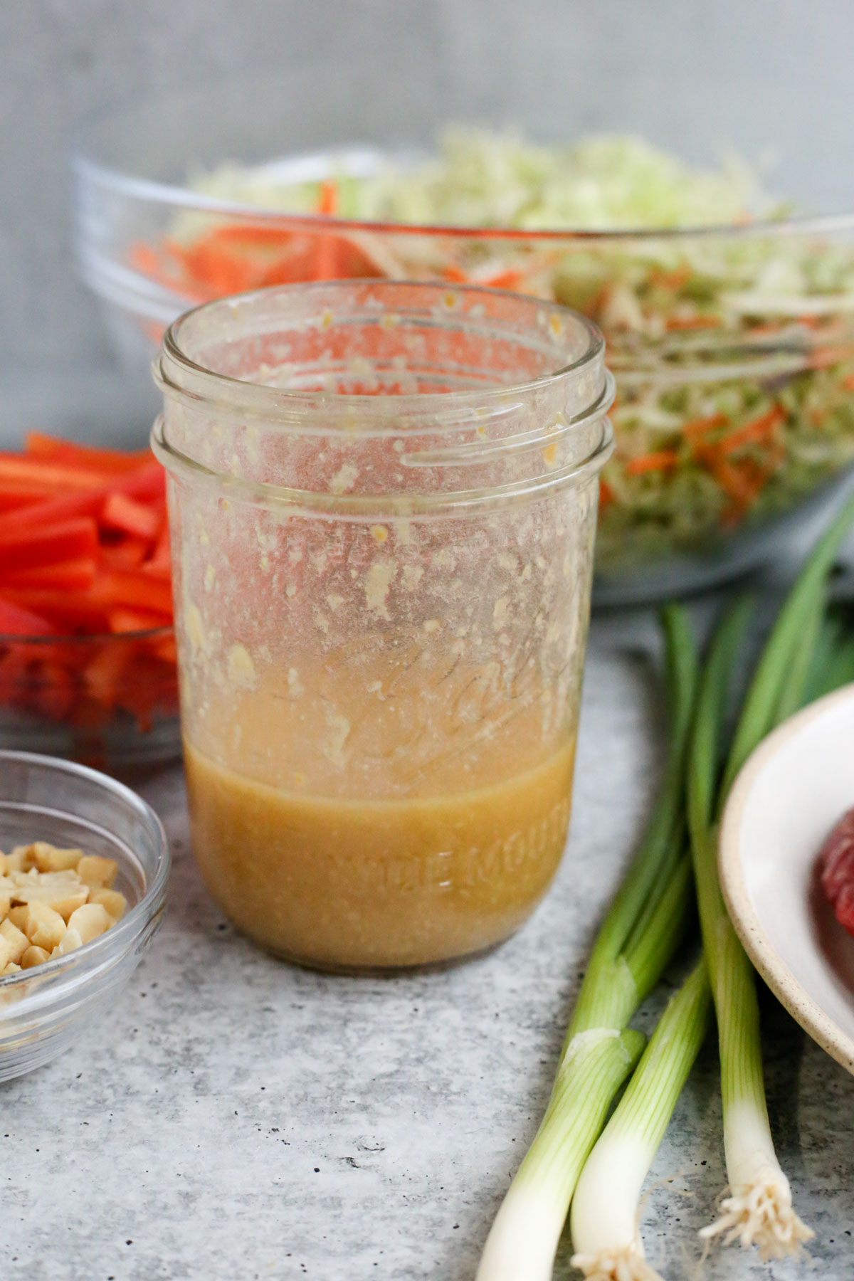 A small glass jar of a chunky salad dressing made using grated ginger and miso, sitting on a cement colored kitchen countertop in from of a large mixing bowl with steak salad ingredients