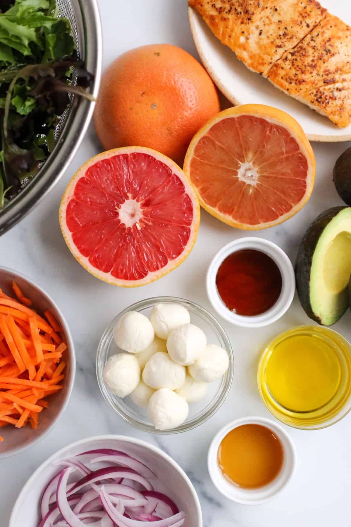 Arrangement of ingredients for a simple grapefruit salad with salmon, set out in small glass ramekins. Includes sliced red onion, olive oil, vinegar, avocado, fresh grapefruit halves, thinly sliced carrots, washed salad greens in a silver metal colander, and a cooked piece of salmon