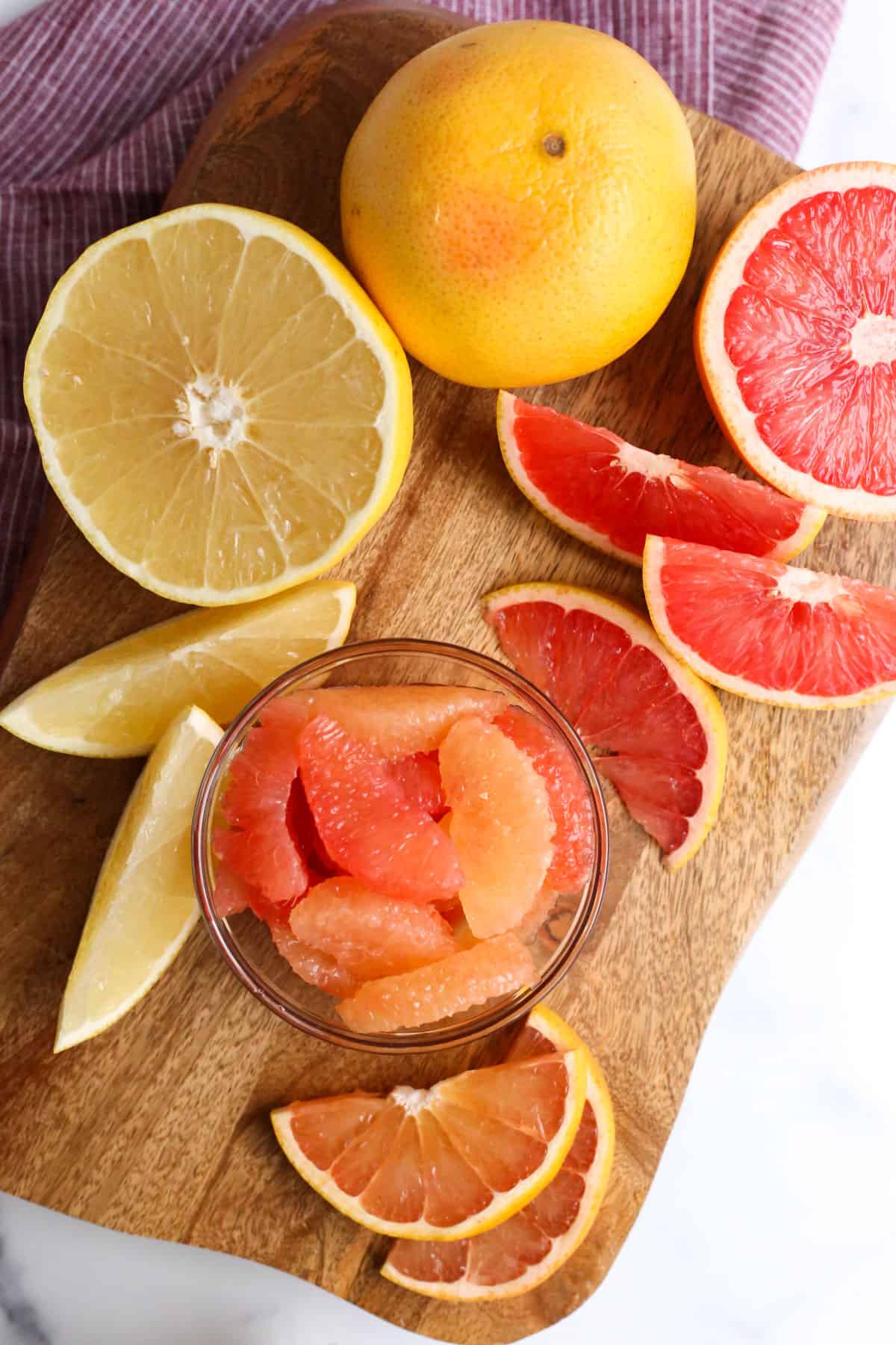 Overhead view of colorful grapefruit segments in a small glass dish, on a wooden cutting board and surround by slices and halves of fresh grapefruit in varying colors