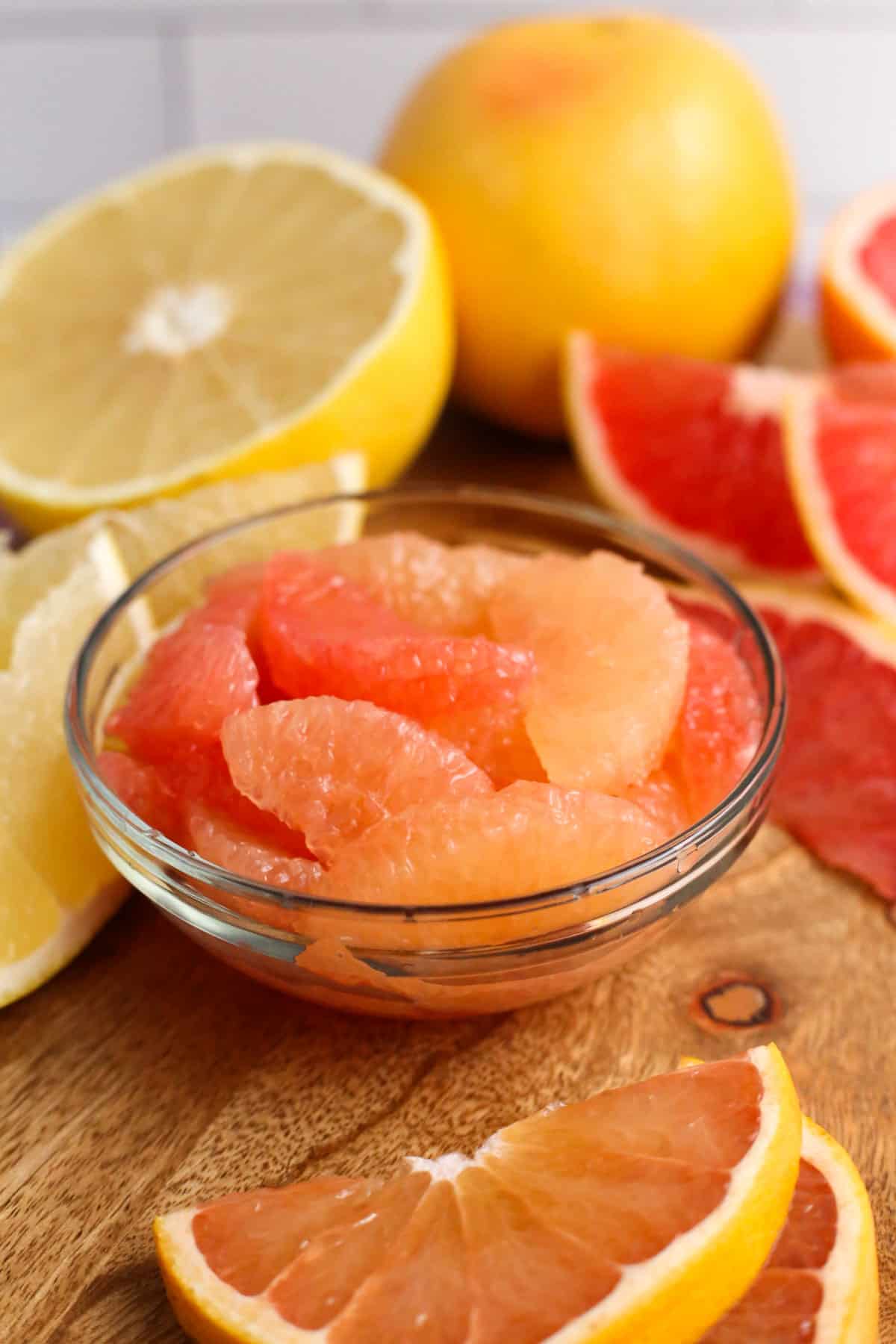 A small glass ramekin with grapefruit segments on a butcher block with several different varieties of grapefruit in the background, ranging from pale yellow to vibrant orange to deep ruby red