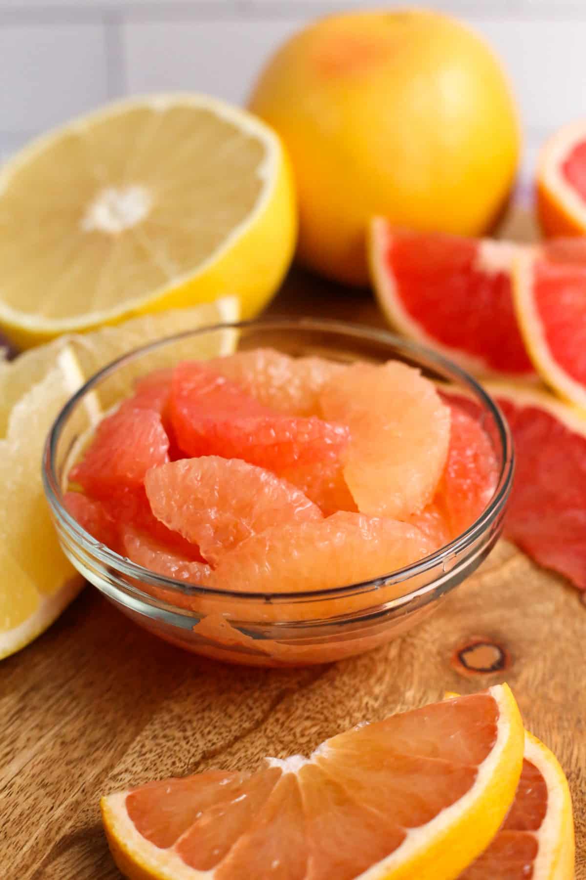 Close up view of supreme grapefruit segments in varying shades of orange and yellow, served in a small glass bowl with sliced grapefruit in the background