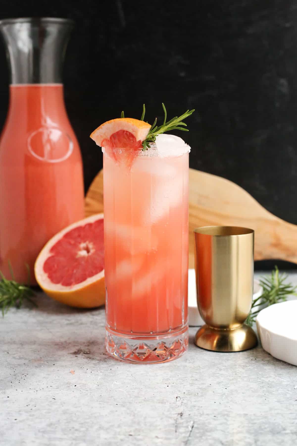 A paloma-style cocktail in a clear highball glass in front of a sliced grapefruit and carafe of grapefruit juice on a kitchen countertop