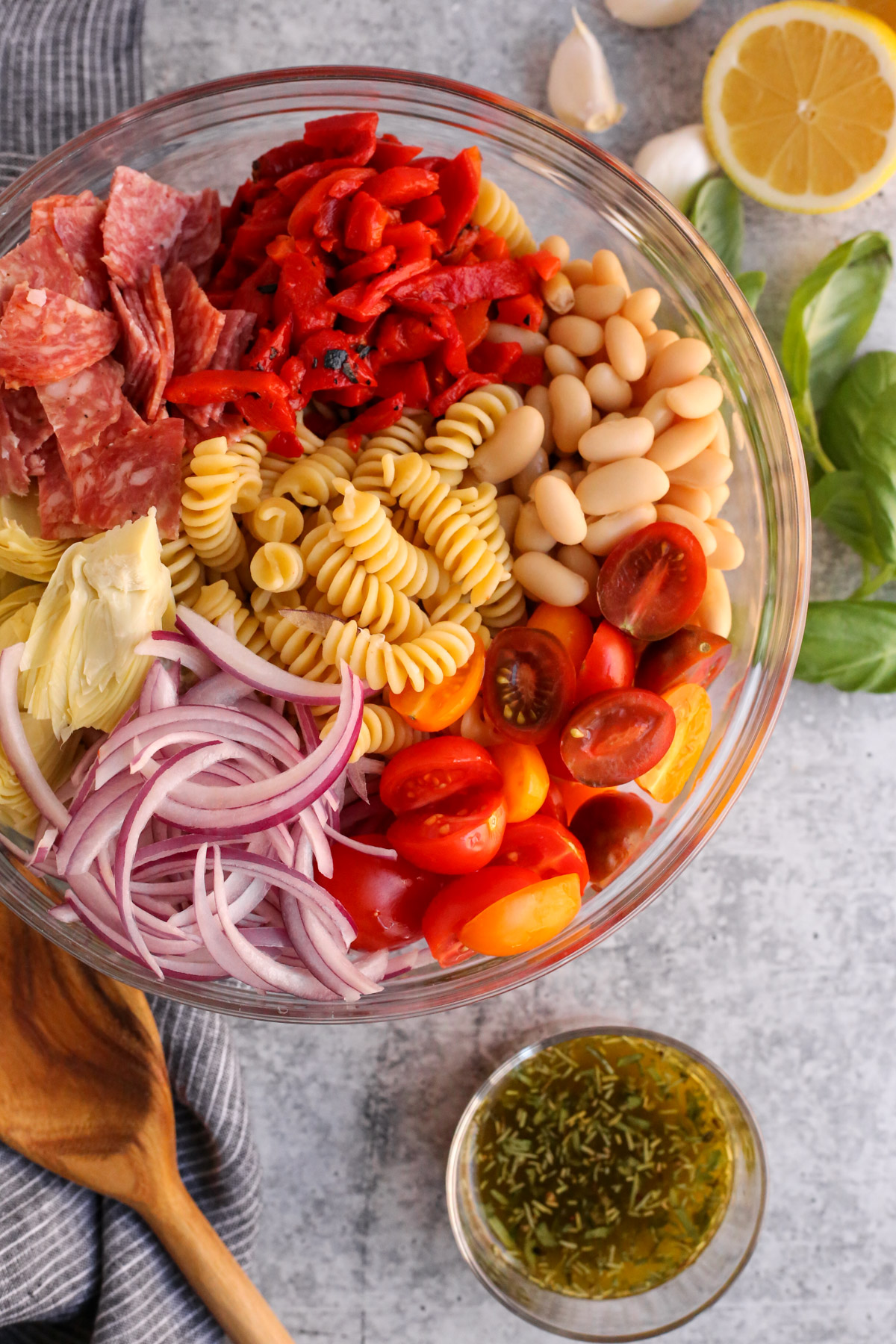 An overhead view of a clear glass mixing bowl containing sliced red onions, artichoke hearts, sliced pepperoni, roasted red peppers, cannellini beans, cherry tomatoes, and cooked pasta, with an Italian-style vinaigrette, fresh basil, lemon, and garlic cloves are displayed around it
